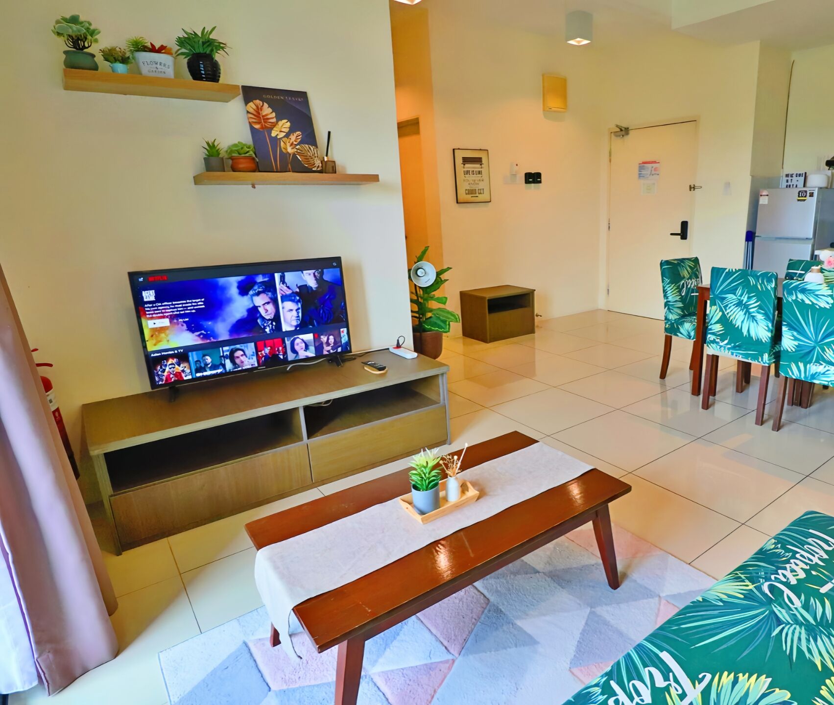 A hotel lobby with a television on a wooden stand, a bench, a small table with a plant, and a dining area with chairs.