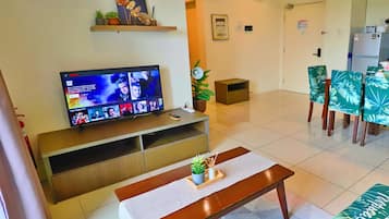 A hotel lobby with a television on a wooden stand, a bench, a small table with a plant, and a dining area with chairs.