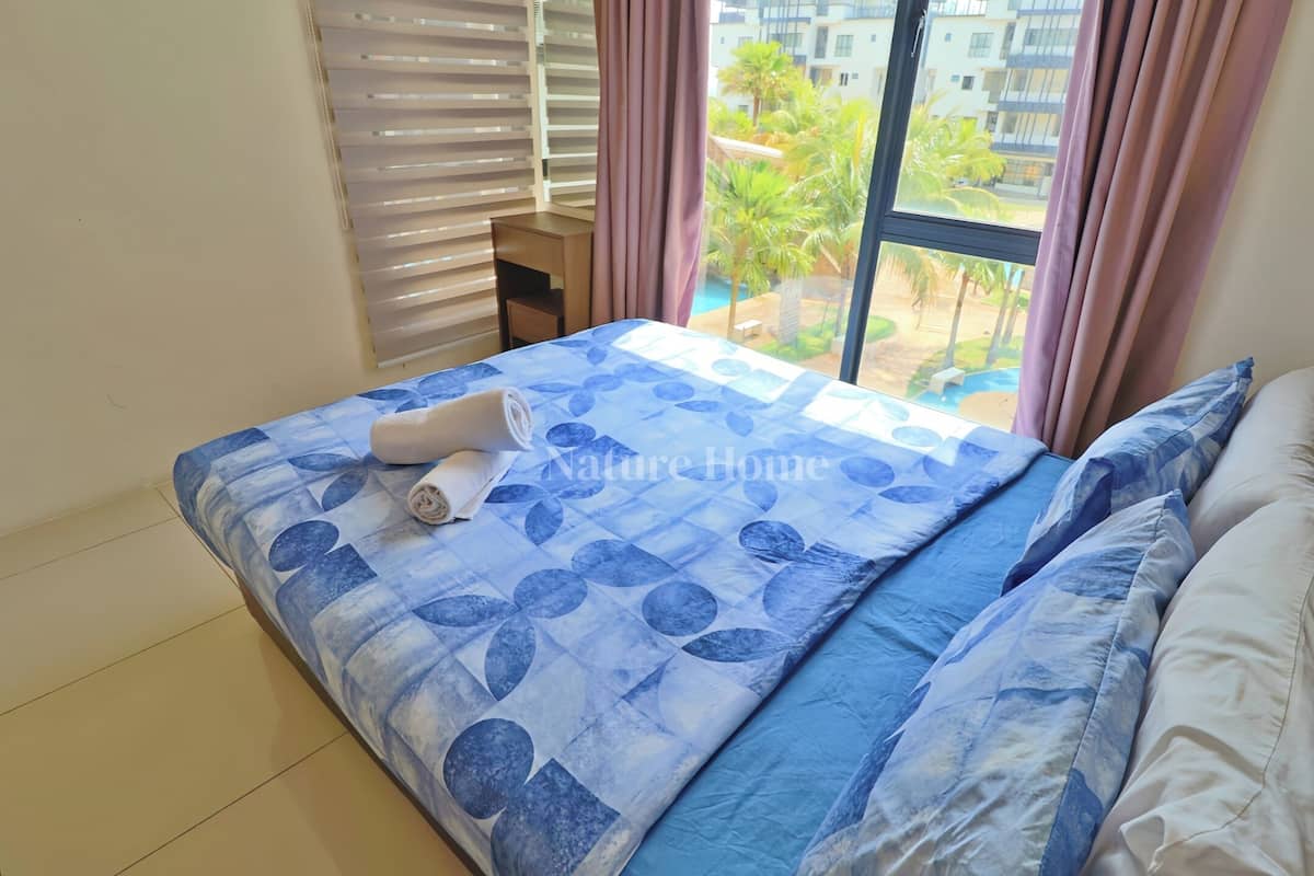 Une chambre avec une grande fenêtre, un lit recouvert d’une courtepointe à motifs bleus, un petit bureau et une vue sur une piscine et la verdure environnante.