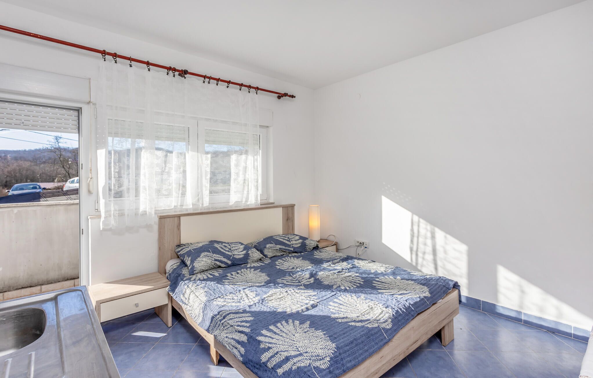 Une chambre à coucher avec un lit, une fenêtre avec des rideaux, une table de chevet avec une lampe, et une vue sur l’extérieur.
