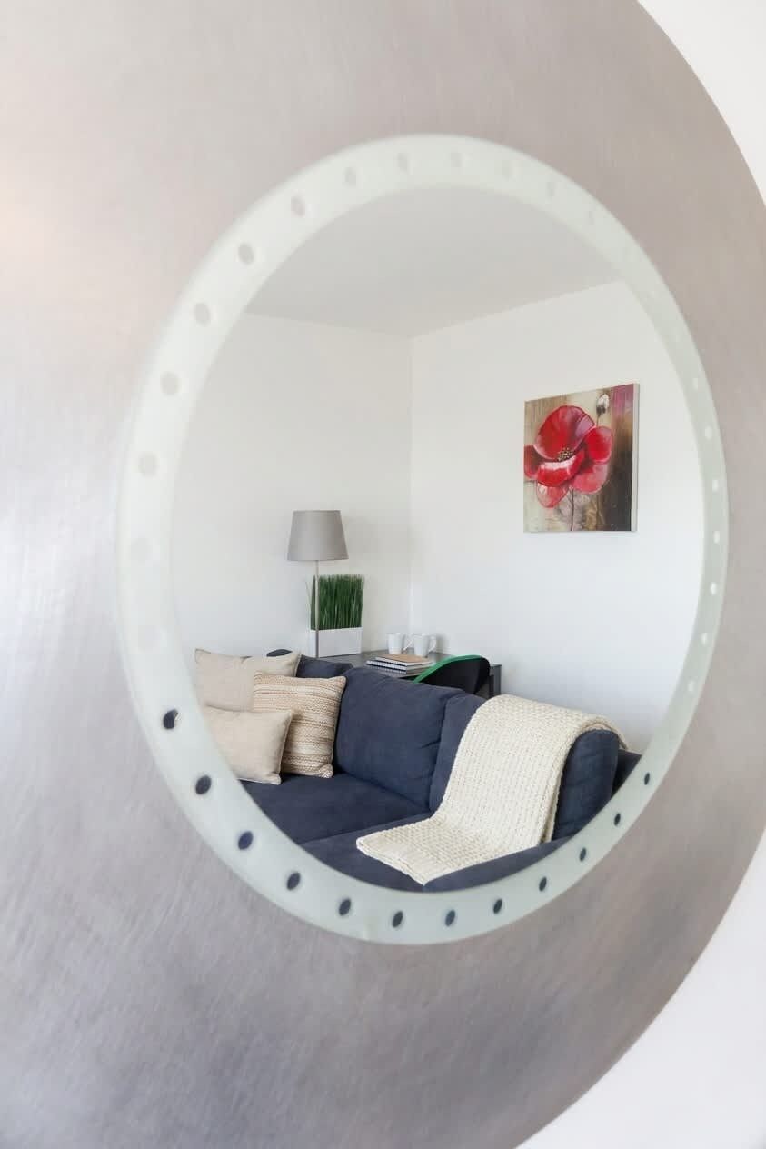 A circular mirror reflecting a living room with a sofa, pillows, a lamp, and a painting.