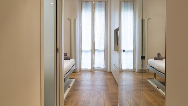 Een moderne slaapkamer met een grote schuifdeur naar een balkon, een bed met matras en kussens, en een inbouwsbank aan het voeteneinde van het bed.
