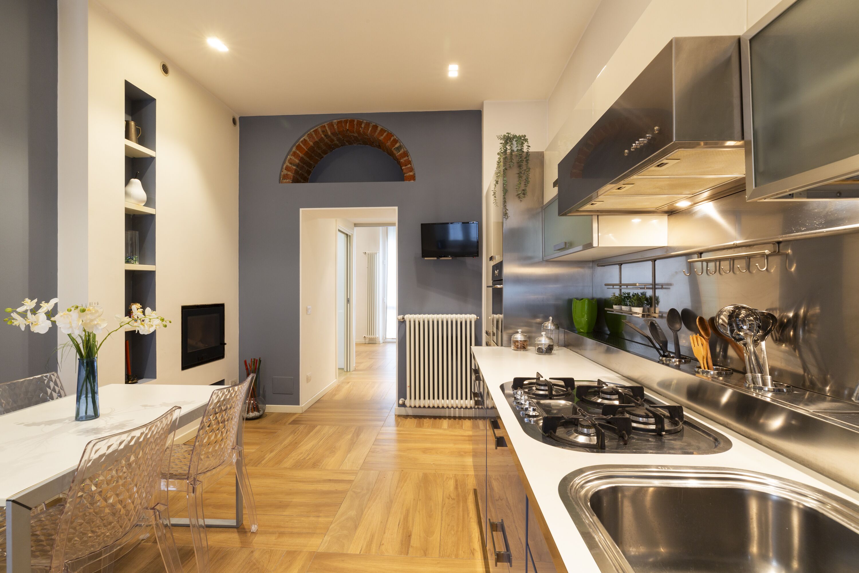 A modern kitchen with a white countertop, stainless steel sink, and a built-in oven. There is a dining area with a white table and chairs, and a brick archway leading to another room.