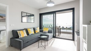 Ein modernes Wohnzimmer mit einer grauen Couch, gelben Kissen, einem kleinen runden Tisch und einer großen Glasschiebetür, die auf einen Balkon mit Meerblick führt.