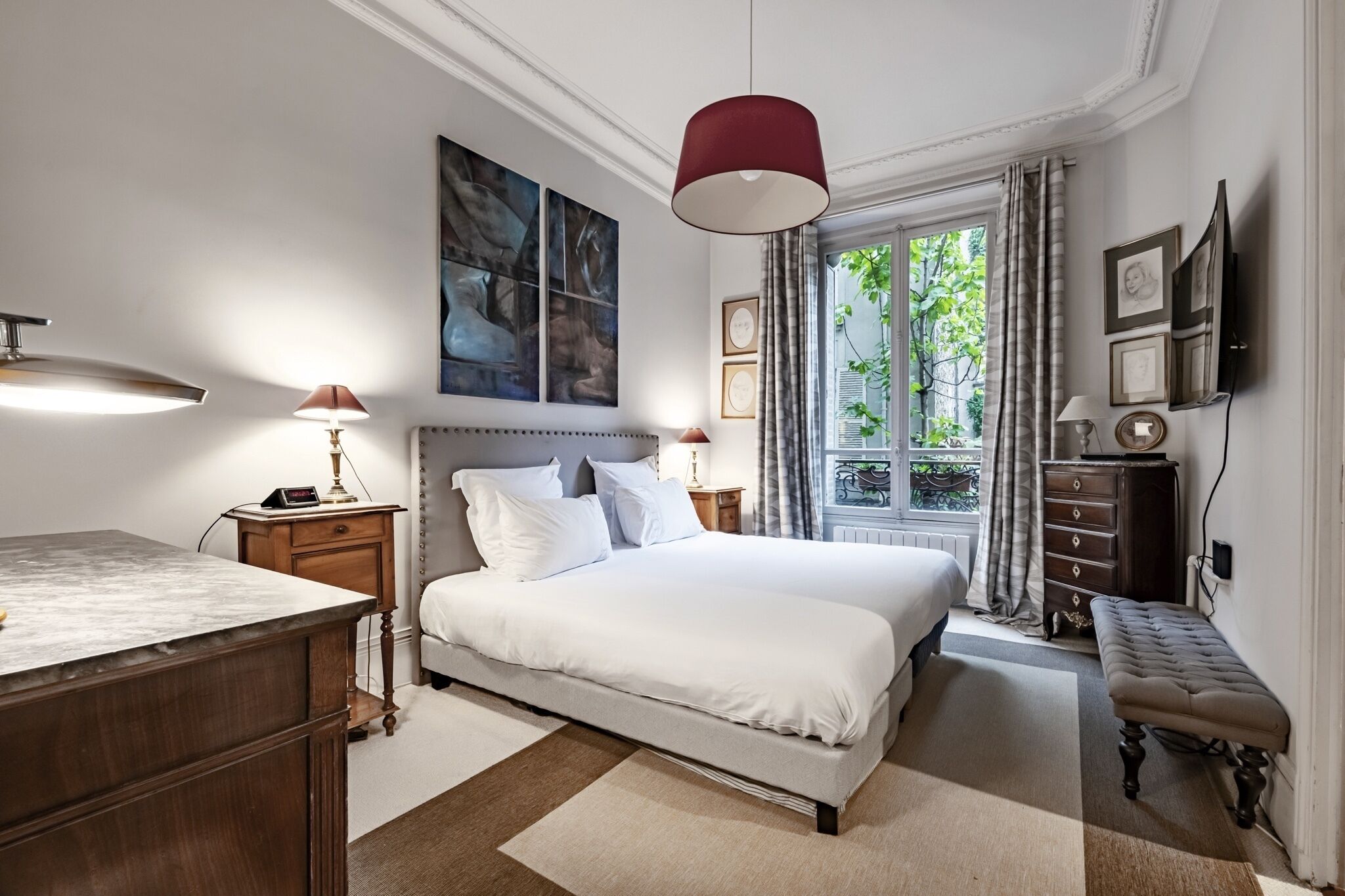 A bedroom with a large bed, bedside tables, a desk, a bench, a window with curtains, and artwork on the walls.