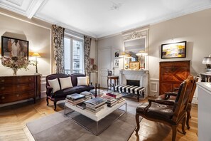 A well-furnished living room with a sofa, armchairs, a coffee table with books, a fireplace, and a large window with curtains. - Epic Romance, by Veeve (Paris)