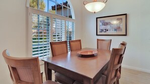 Un coin repas avec une table et des chaises en bois, une grande fenêtre avec des stores et un tableau encadré au mur.
