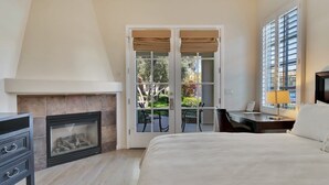 A bedroom with a fireplace, a bed, a desk with a lamp, and a view through a glass door to an outdoor area. - The Palm Retreat B | Studio Legacy Villa (La Quinta)