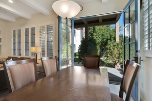 A dining area with a large table and chairs, a lamp, and a view of a garden through open doors. - Desert Haven A | 1BR Legacy Villa (La Quinta)