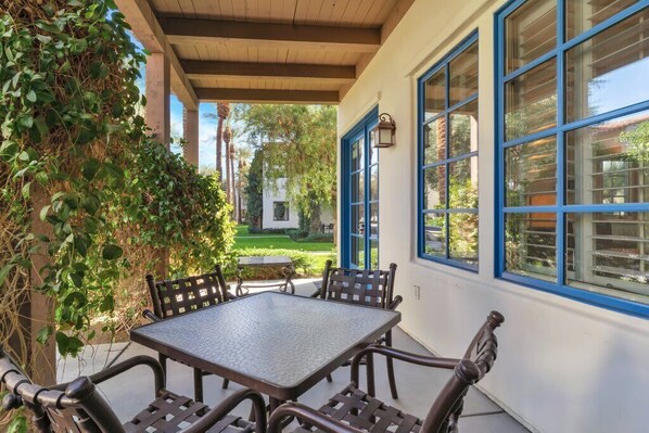 Un patio exterior con una mesa y sillas, rodeado por un muro blanco con ventanas de marco azul.