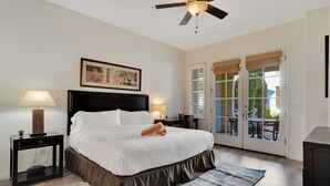 Habitación de hotel con cama, ventilador de techo, un cuadro, una mesita de noche, lámpara, alfombra y puerta de vidrio corredera que da a un área exterior.