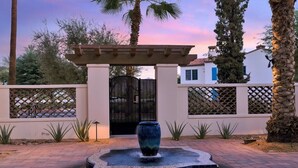 A gated entrance with a water feature in the foreground, palm trees, and a building with a blue door in the background. - Desert Haven C | Studio Legacy Villa (La Quinta)