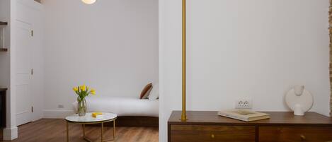 A modern room with a wooden floor, a round table with a vase of yellow flowers, a bed with white bedding, and a desk with a book and a vase.