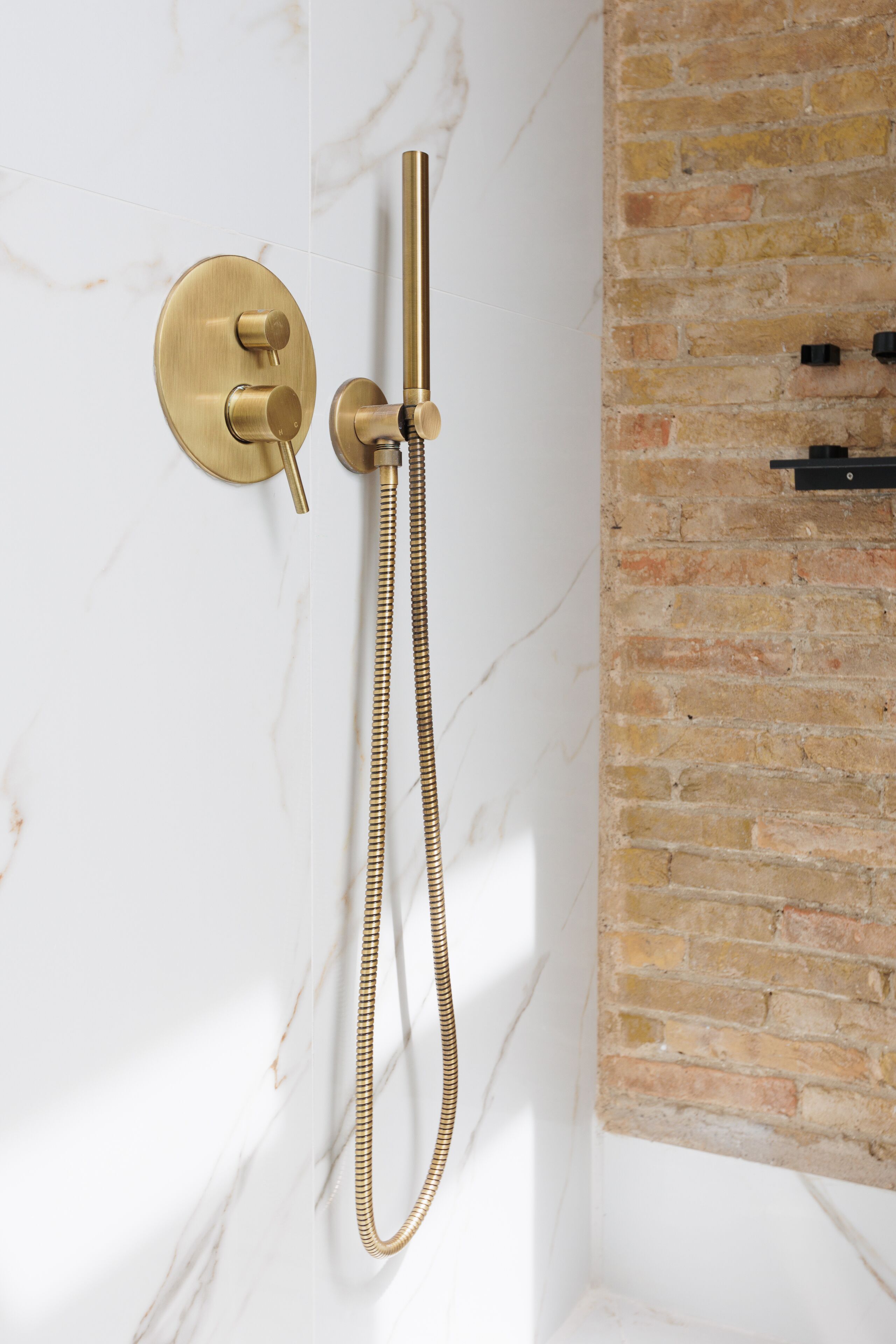 A modern bathroom with a gold showerhead and hose, marble walls, and a brick accent wall.