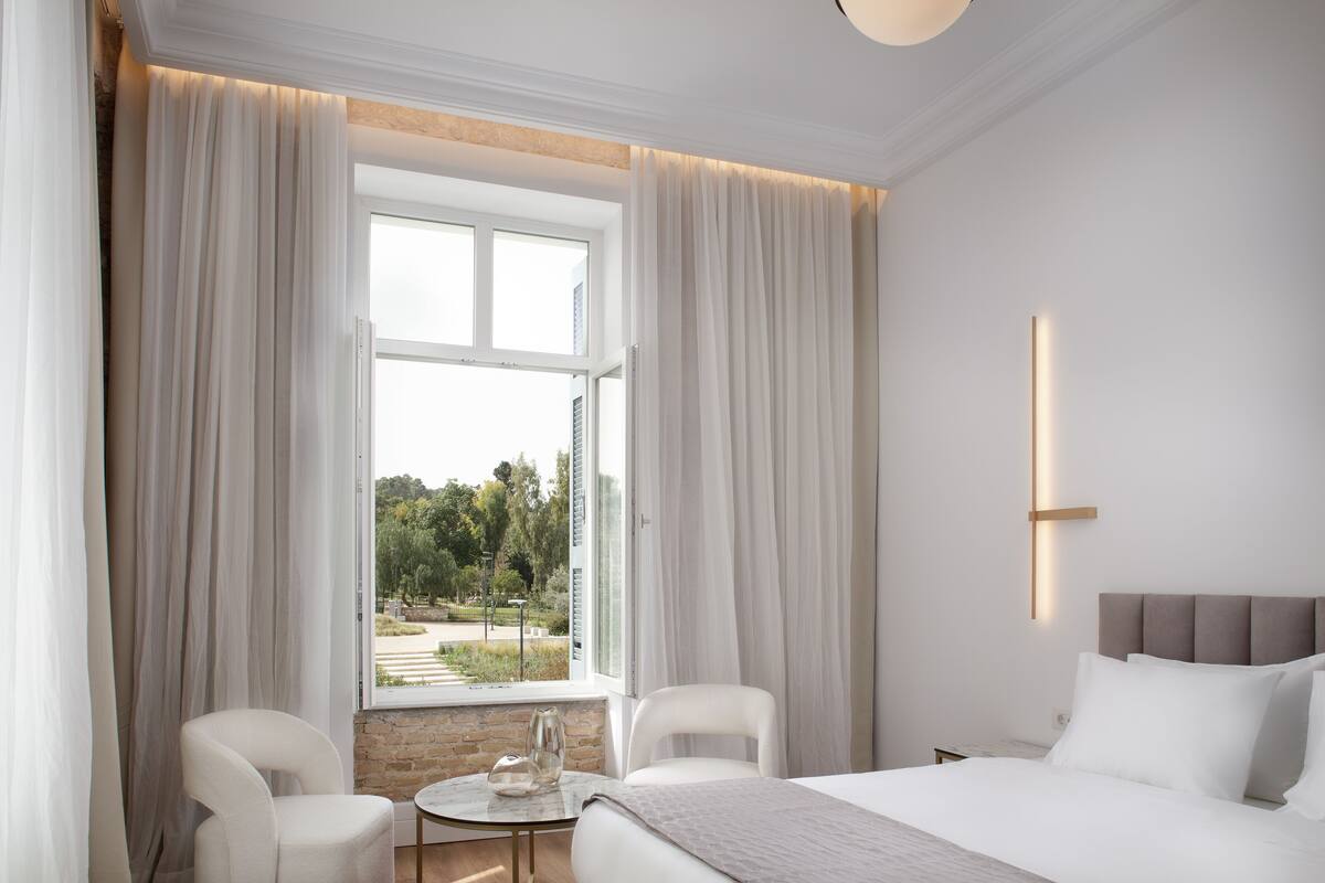A modern hotel room with a large bed, two chairs, a small table, and a view of the outdoors through a large window.
