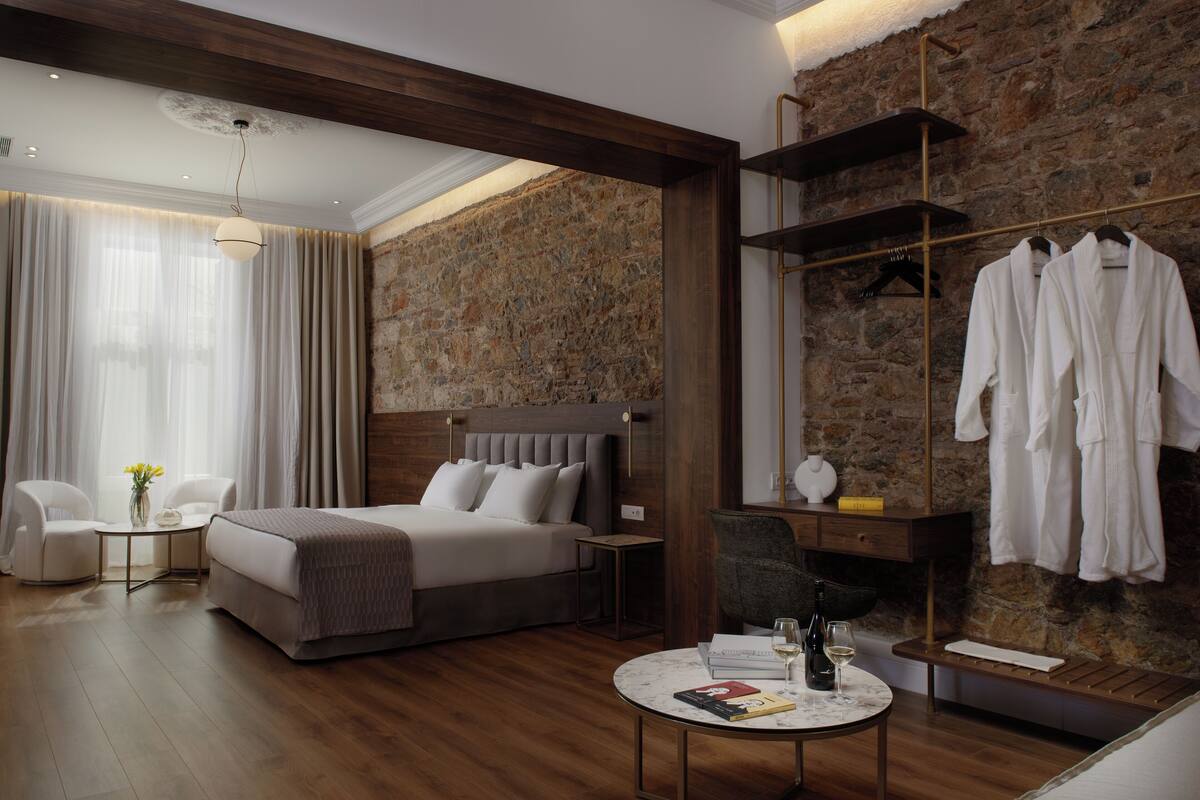 A modern hotel room with a large bed, a round table with a wine bottle, a small table with a vase, and a robe rack.