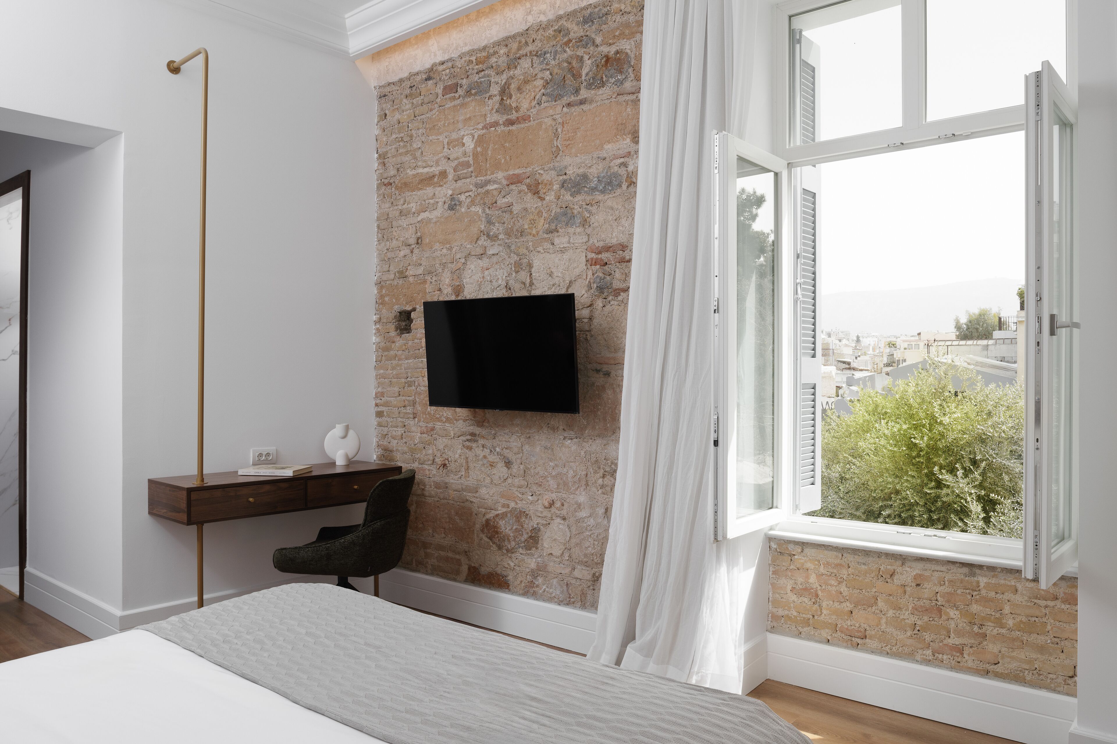 A bedroom with a brick wall, a flat-screen TV, a desk, a chair, and a large window with a view of buildings.