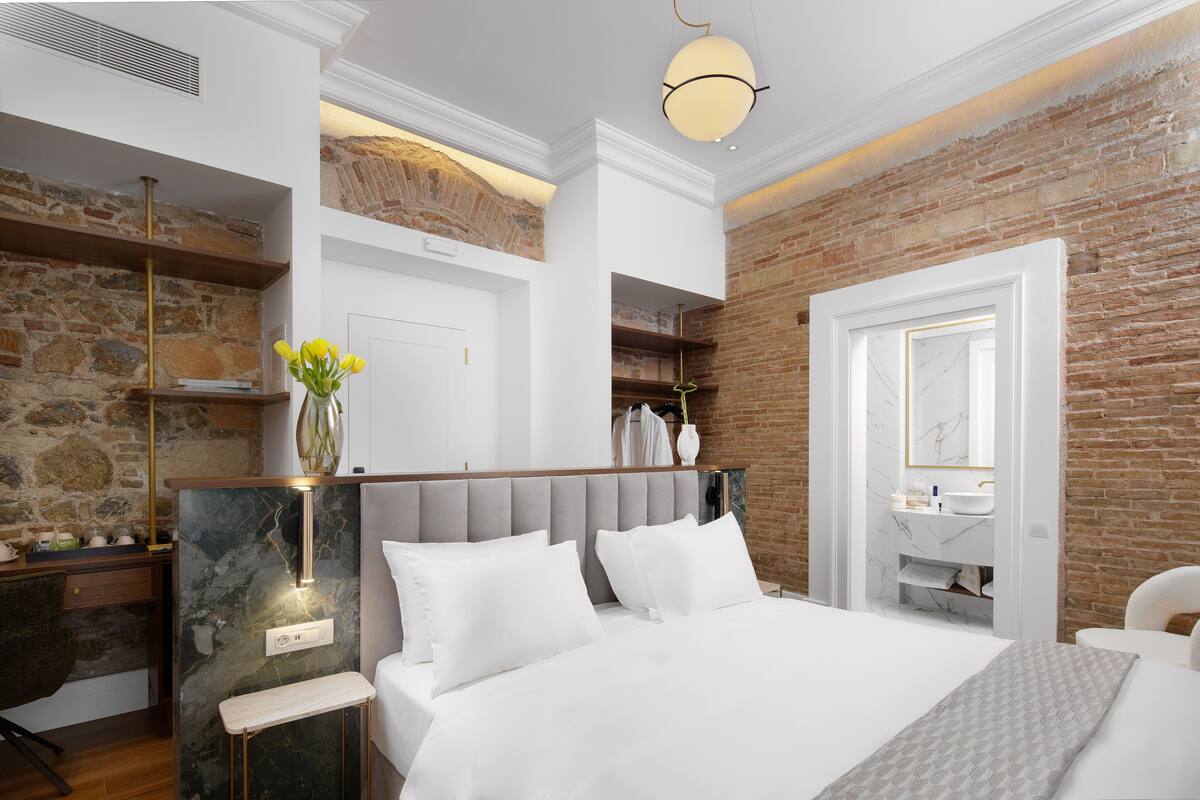 A modern bedroom with a bed, a nightstand, a desk, and a bathroom visible through an open door.