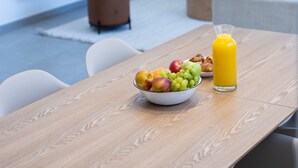 Un soggiorno moderno con un tavolo da pranzo in legno, una ciotola di frutta e un bicchiere di succo d'arancia. Una TV a schermo piatto è montata a parete, e si intravede un pouf marrone.