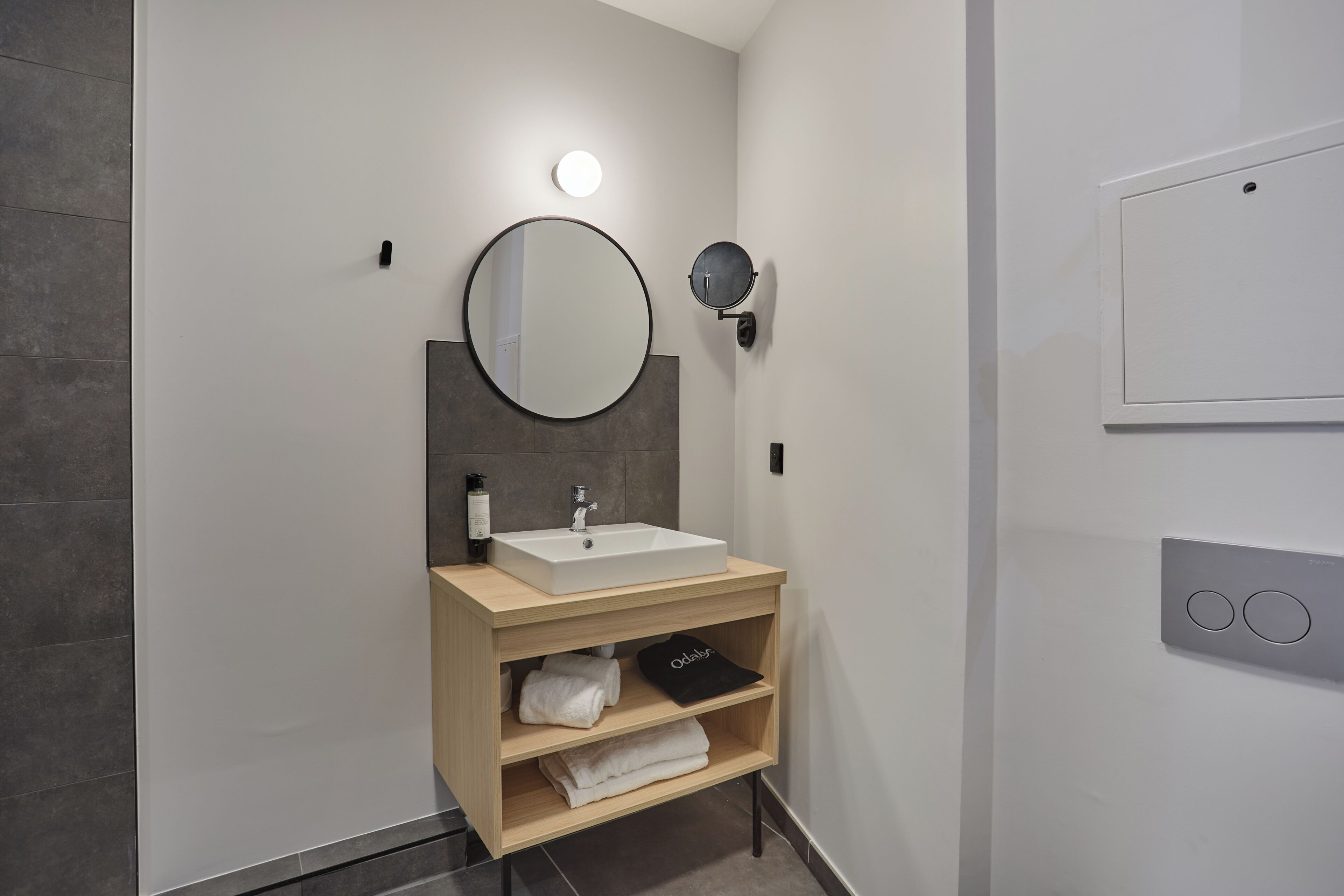 A modern bathroom with a white sink, a round mirror, a towel shelf, and a wall-mounted light.