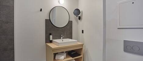 A modern bathroom with a white sink, a round mirror, a towel shelf, and a wall-mounted light.