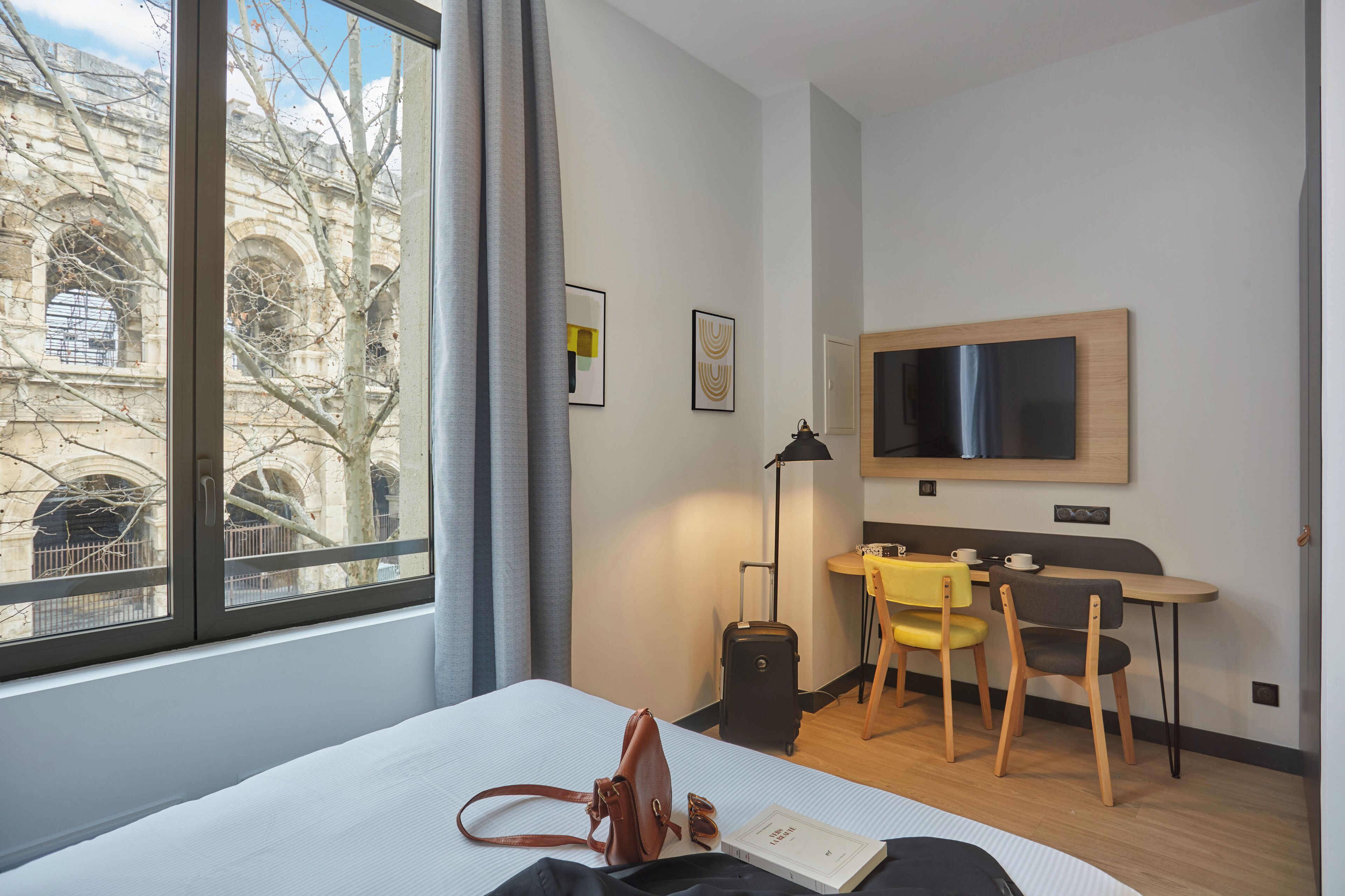 Ein Hotelzimmer mit Bett, Schreibtisch, Stuhl, Koffer und Fernseher an der Wand.