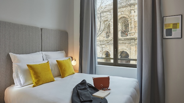 A hotel room with a bed, pillows, a book, a bag, and a window with a view of a building.