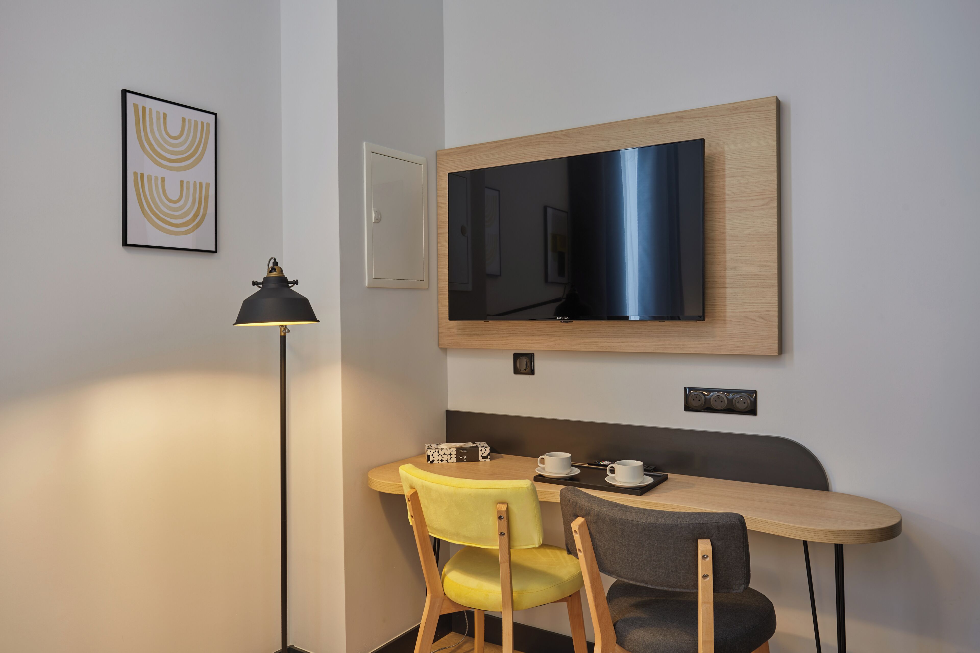 Et moderne værelse med et fjernsyn ophængt på væggen, et skrivebord med to stole, en gulvlampe og et indrammet kunstværk.