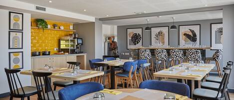 A modern dining area with multiple tables and chairs, a kitchen area with a yellow backsplash, and various framed artworks on the walls.