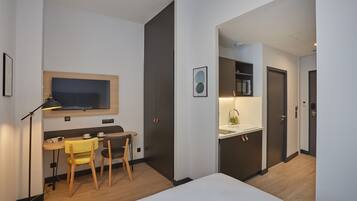 Una habitación de hotel moderna con cama, escritorio con silla, una pequeña mesa con silla, televisor montado en la pared y un área de kitchenette.