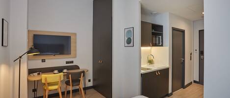 A modern hotel room with a bed, a desk with a chair, a small table with a chair, a TV mounted on the wall, and a kitchenette area.