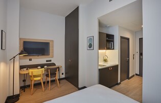 Una habitación de hotel moderna con cama, escritorio con silla, una pequeña mesa con silla, televisor montado en la pared y un área de kitchenette.