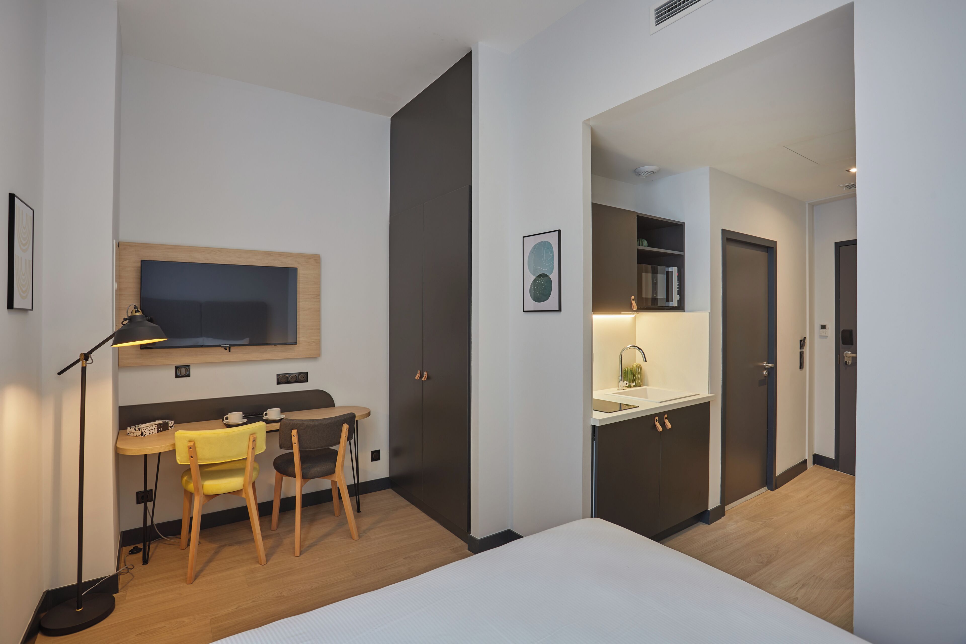 Une chambre d’hôtel moderne équipée d’un lit, d’un bureau avec des chaises, d’une télévision et d’une kitchenette.