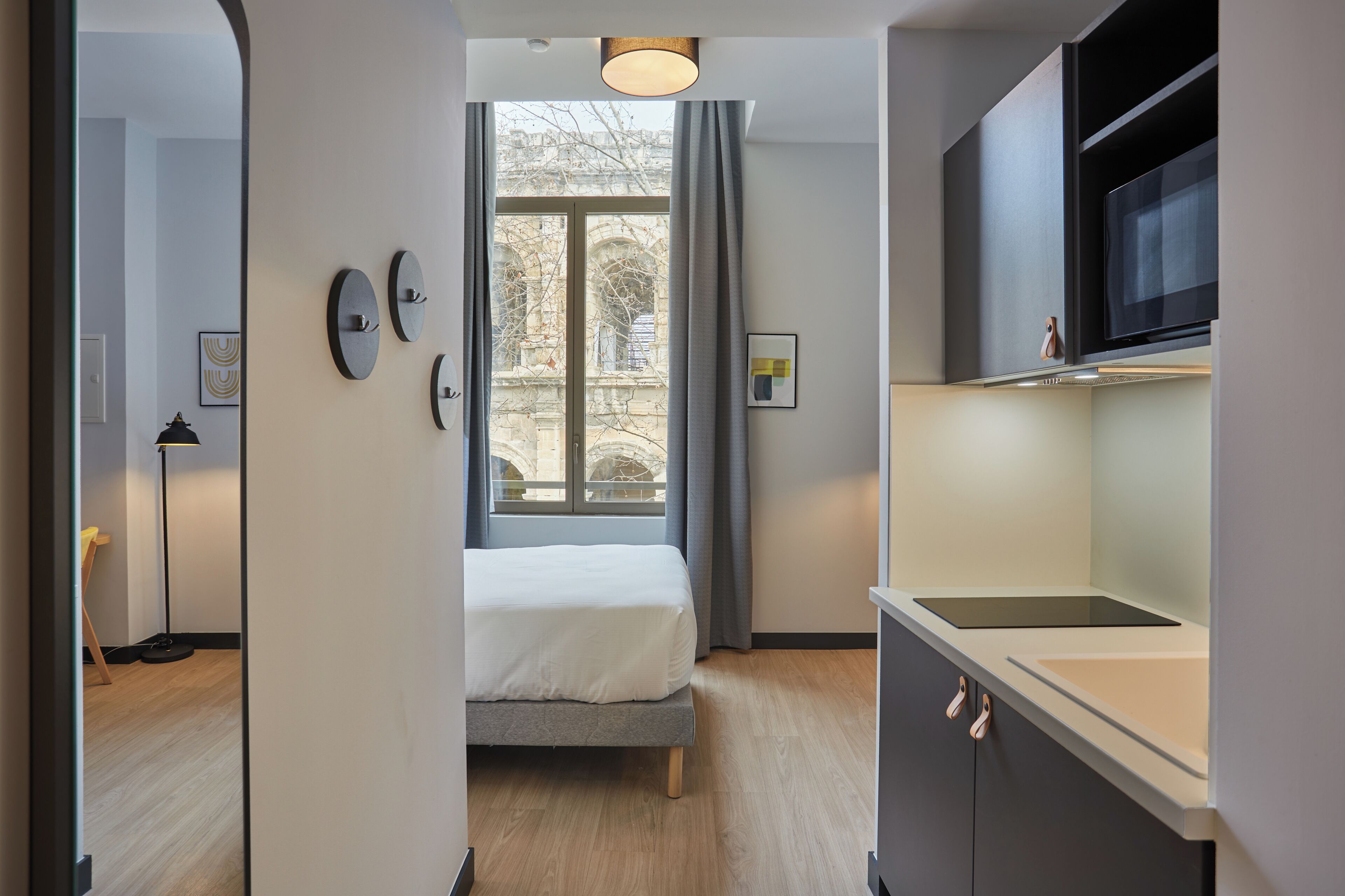 Une chambre moderne comprenant un lit, une fenêtre avec des rideaux, un coin cuisine équipé d’un four à micro-ondes et une porte donnant sur une autre pièce.