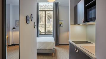 Une chambre moderne comprenant un lit, une fenêtre avec des rideaux, un coin cuisine équipé d’un four à micro-ondes et une porte donnant sur une autre pièce.