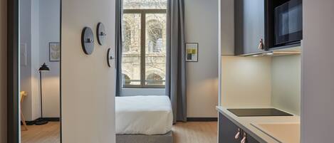 Una habitación moderna con cama, ventana con cortinas, espacio de cocina con microondas y puerta que da a otra habitación.