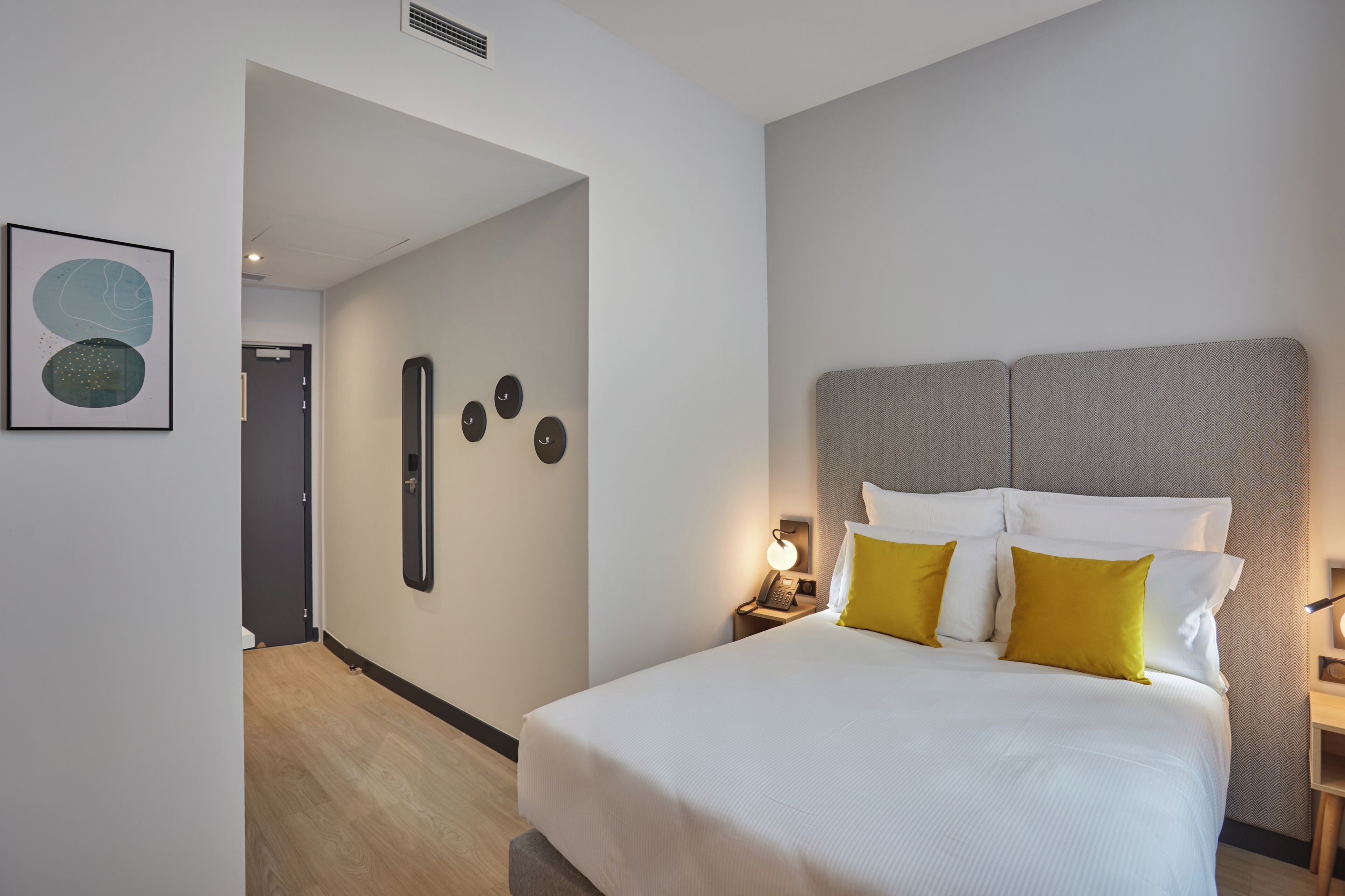 A hotel room with a bed, bedside table, wall clock, and a door leading to another room.