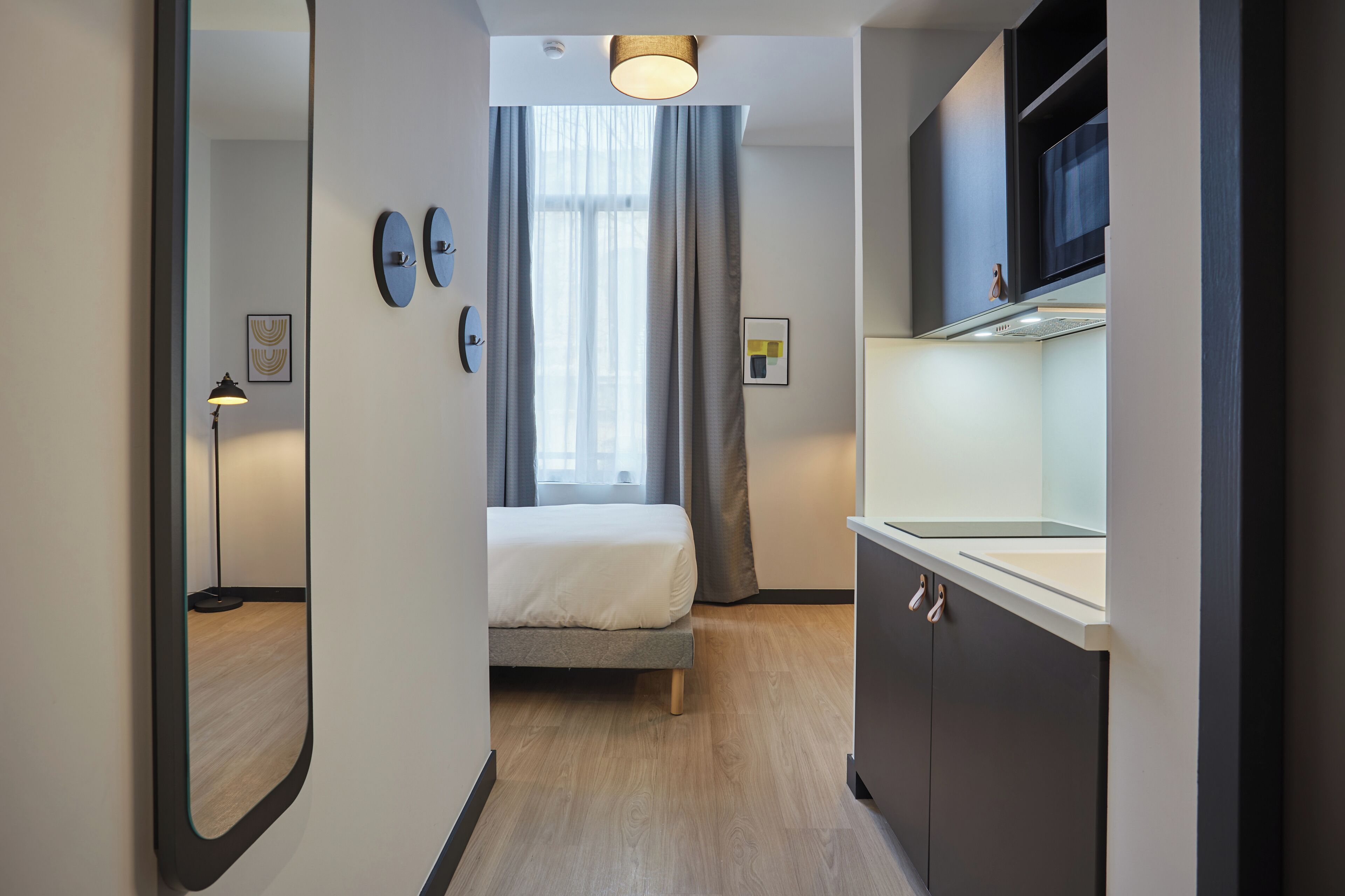 A modern hotel room with a bed, a mirror, a sink, and a window with curtains.