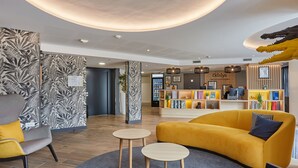 A modern reception area with a yellow sofa, two round tables, and a bookshelf with various items. - Appart'hôtel Odalys City - Nîmes Arènes - Palais des Congrès (Nîmes)