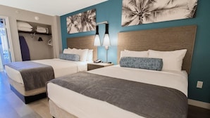 Habitación de hotel con dos camas, una mesita de noche con lámpara y un cuadro con motivos de hojas de palmera.