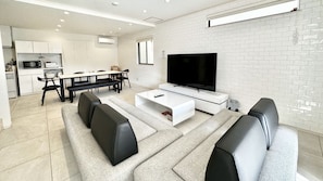 Una sala moderna con un televisor de pantalla plana, un comedor con un banco y una cocina al fondo.