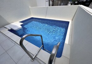 Un pequeño jacuzzi rectangular con una escalera metálica, rodeado de paredes blancas y un piso de baldosas.