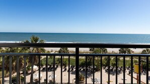 Vista a la playa desde un balcón con barandilla, palmeras y sillas de playa.