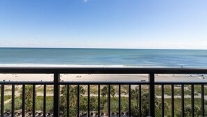 Vista a la playa desde un balcón con barandilla, palmeras y cielo despejado.