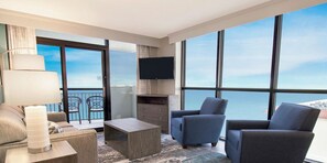A modern hotel room with a large balcony, a flat-screen TV, a sofa, two armchairs, and a coffee table.