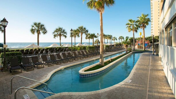 A pool area with lounge chairs, palm trees, and a view of the ocean.