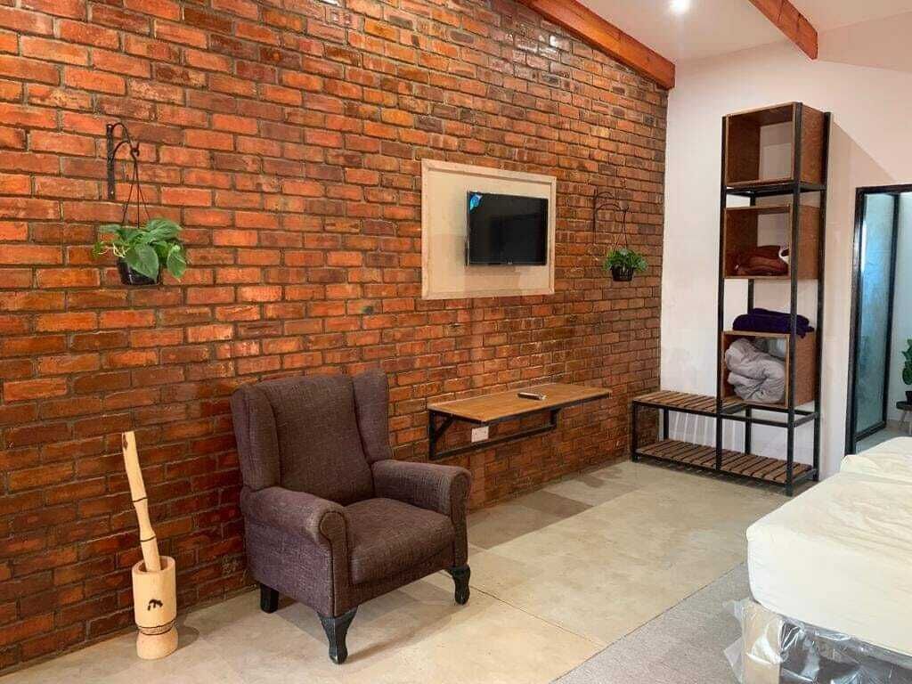 Una habitación con pared de ladrillo, un televisor montado, una mesa de madera, una silla y una cama con colchón en la esquina.
