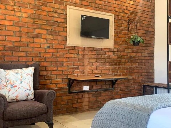 A room with a brick wall, a mounted TV, a small table, a chair, and a bed.