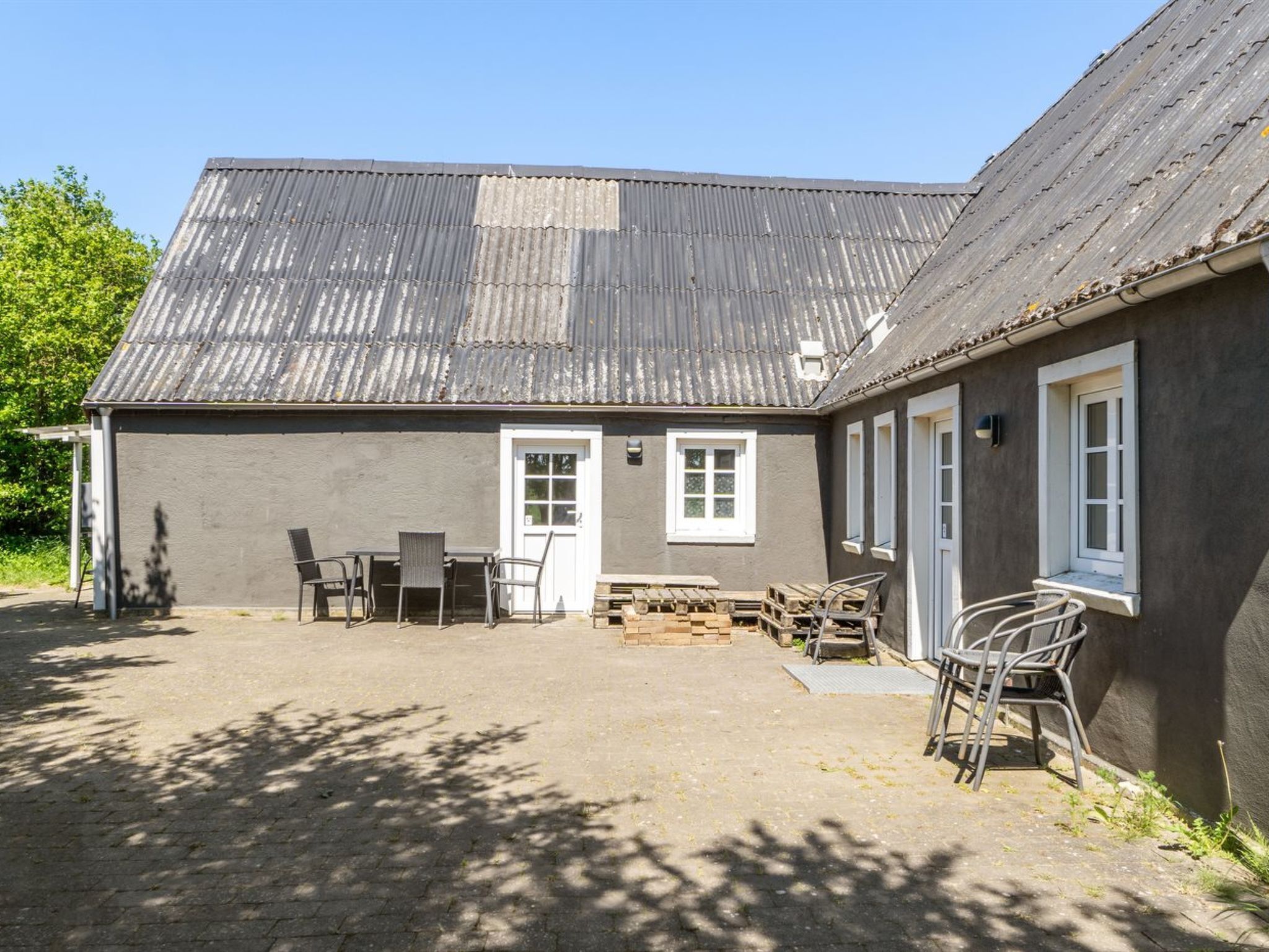 Rakennuksessa on aaltopeltikatto, pieni patio, jossa on pöytä ja tuoleja, sekä ikkunoilla varustettu ovi.