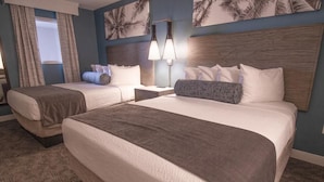 Habitación de hotel con dos camas, una mesita de noche, una lámpara y un cuadro con palmeras en la pared.